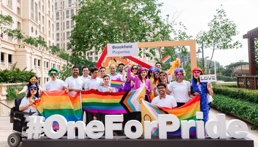 Group of people celebrating Pride at Downtown Powai, holding rainbow flags and standing behind a #OneForPride sign